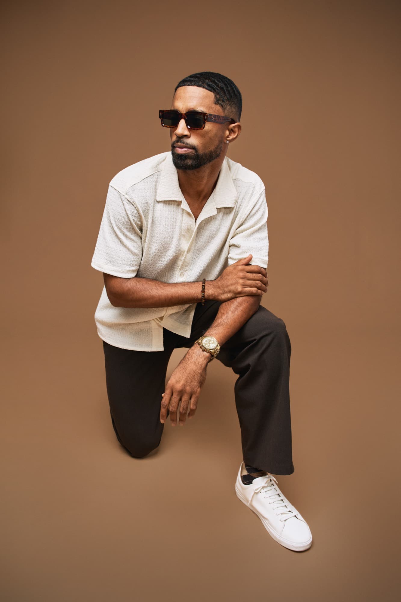 Abyon Ausar crouched in a cream camp shirt and sunglasses against a warm backdrop