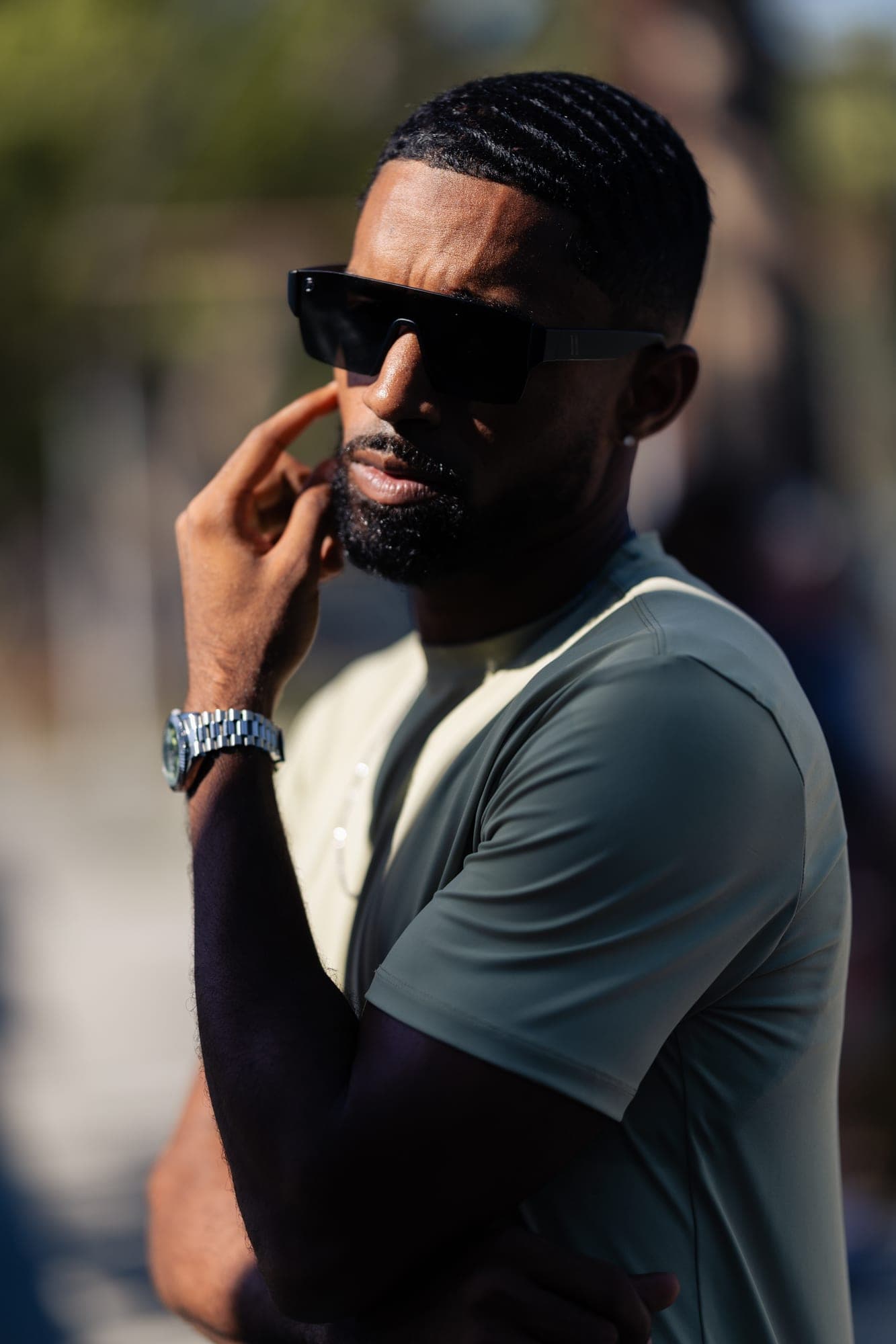 Abyon Ausar in sunglasses and an olive tee, hand raised to his face, on a sunlit street